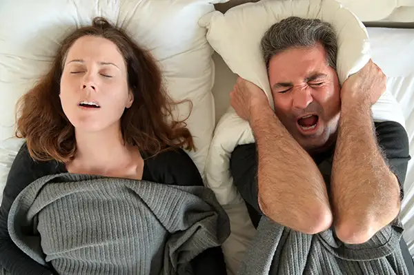 Woman snoring loudly in bed while her partner holds a pillow over his ears, struggling to sleep.